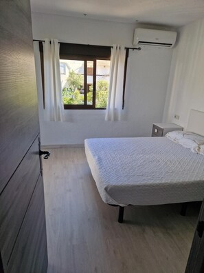 Ein Schlafzimmer mit einem Bett, einem Fenster mit Vorhängen, einer Klimaanlage und einem kleinen Nachttisch.