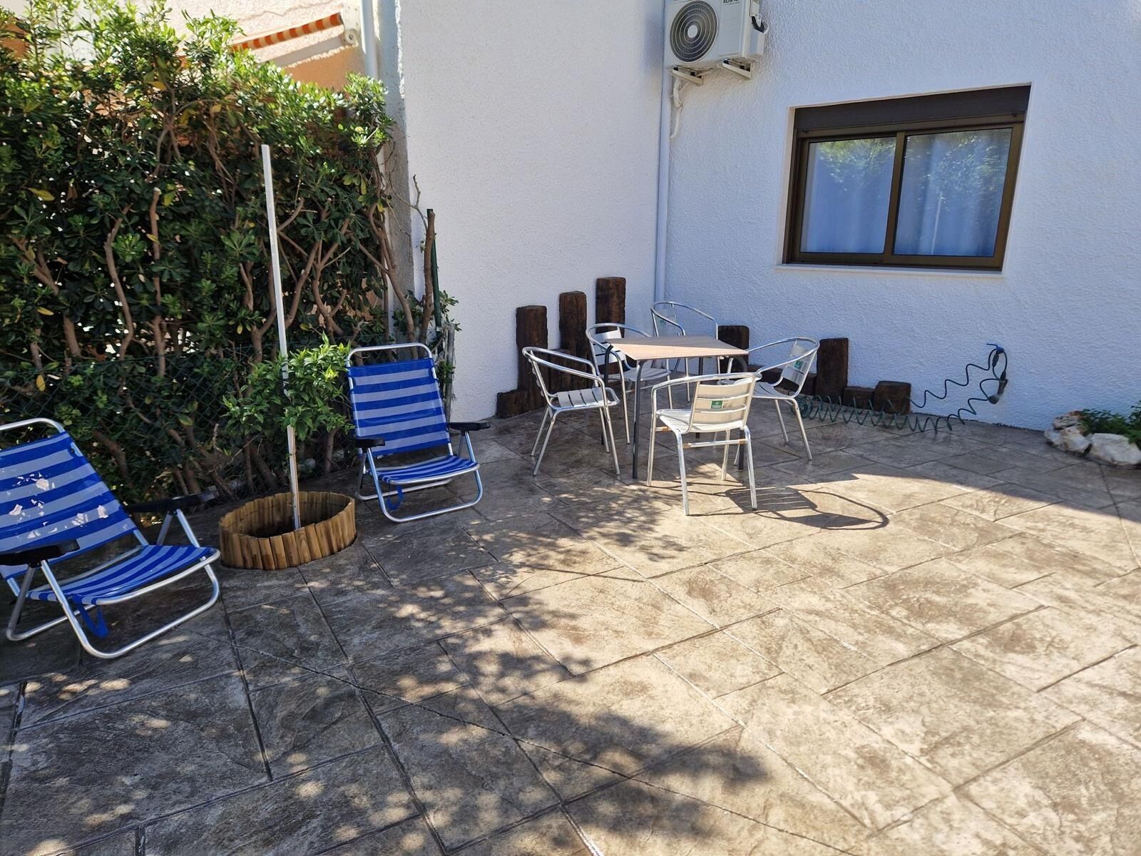 En udendørs terrasse med et bord, stole, en lille træplantekasse og en hvid væg med et vindue.