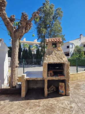 Ein steinernes Grillhaus im Freien mit Schornstein, einem Baum und einem eingezäunten Bereich mit Gebäuden im Hintergrund.