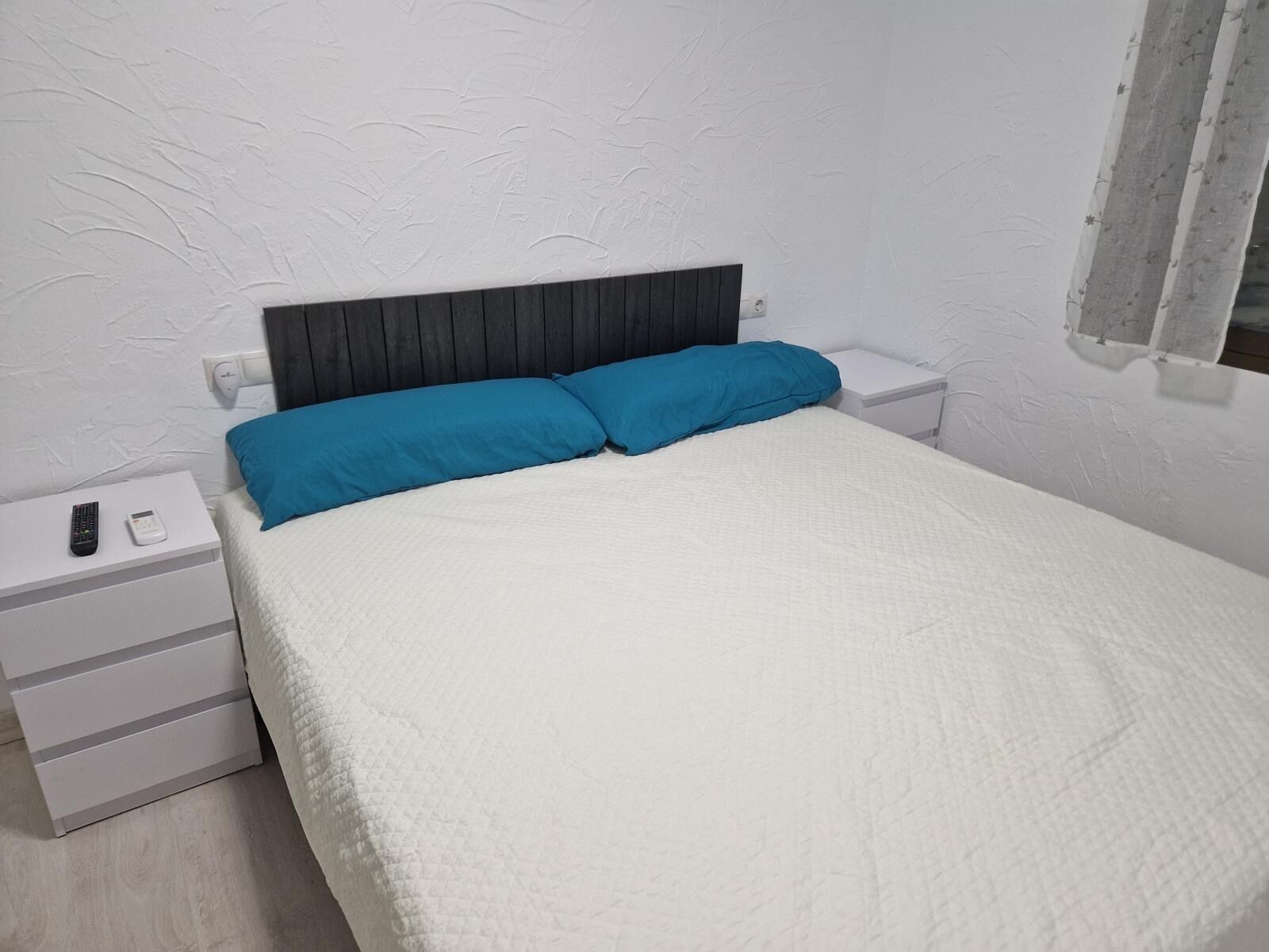 A bedroom with a bed, two pillows, a nightstand, a remote, and a wall socket.