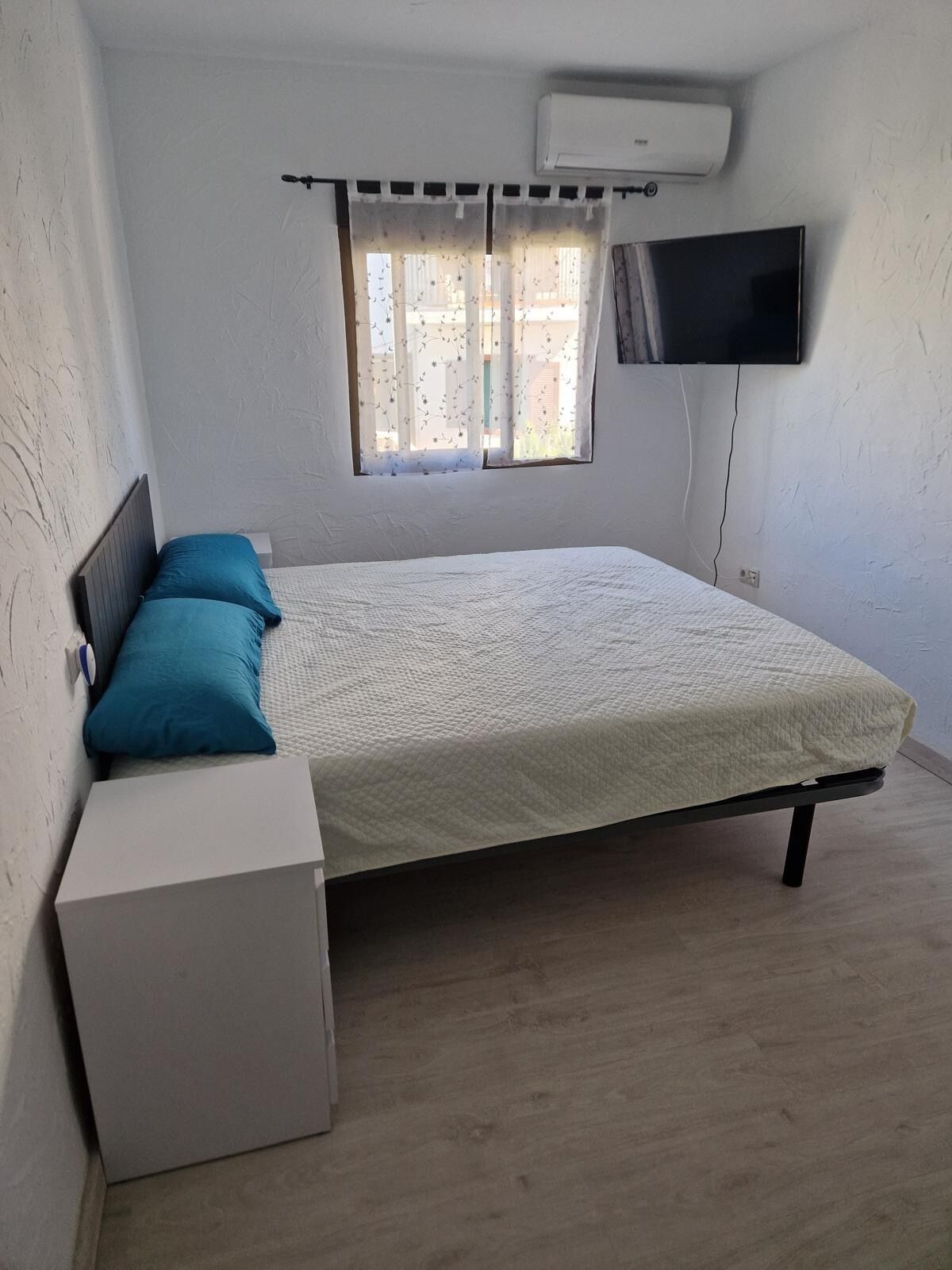 Une chambre à coucher comprenant un lit, une petite table de chevet, un téléviseur fixé au mur et une fenêtre avec des rideaux.
