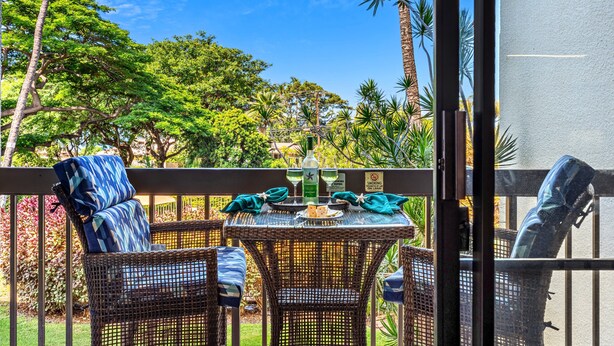A balcony with wicker furniture, a table set with a bottle and glasses, and a view of tropical trees. - Tropical Retreat, steps from Charlie Young Beach (Kihei)