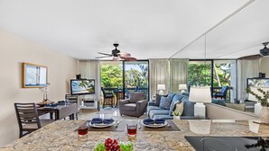 A spacious living area with a marble island, seating, a television, and a view of greenery outside. - Tropical Retreat, steps from Charlie Young Beach (Kihei)