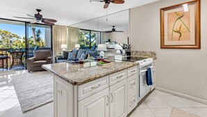 A modern kitchen with a large island, a dining area with a sofa, and a balcony with a view of palm trees. - Tropical Retreat, steps from Charlie Young Beach (Kihei)