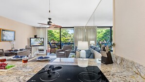A modern kitchen with a built-in stove, dining area with a table and chairs, and a living area with a sofa and a TV. - Tropical Retreat, steps from Charlie Young Beach (Kihei)