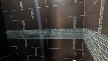 A bathroom with dark tiled walls, a small shelf with a green bottle, and a textured floor.