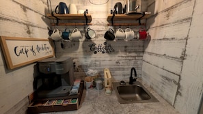 Una kitchenette pequeña con cafetera, tazas, un lavabo y un cartel enmarcado.