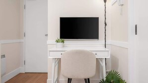 Ein weißer Schreibtisch mit einem schwarzen Monitor, einem Stuhl, einer Topfpflanze und einer Stehlampe.