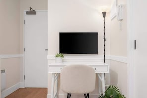 Ein weißer Schreibtisch mit einem schwarzen Monitor, einem Stuhl, einer Topfpflanze und einer Stehlampe.
