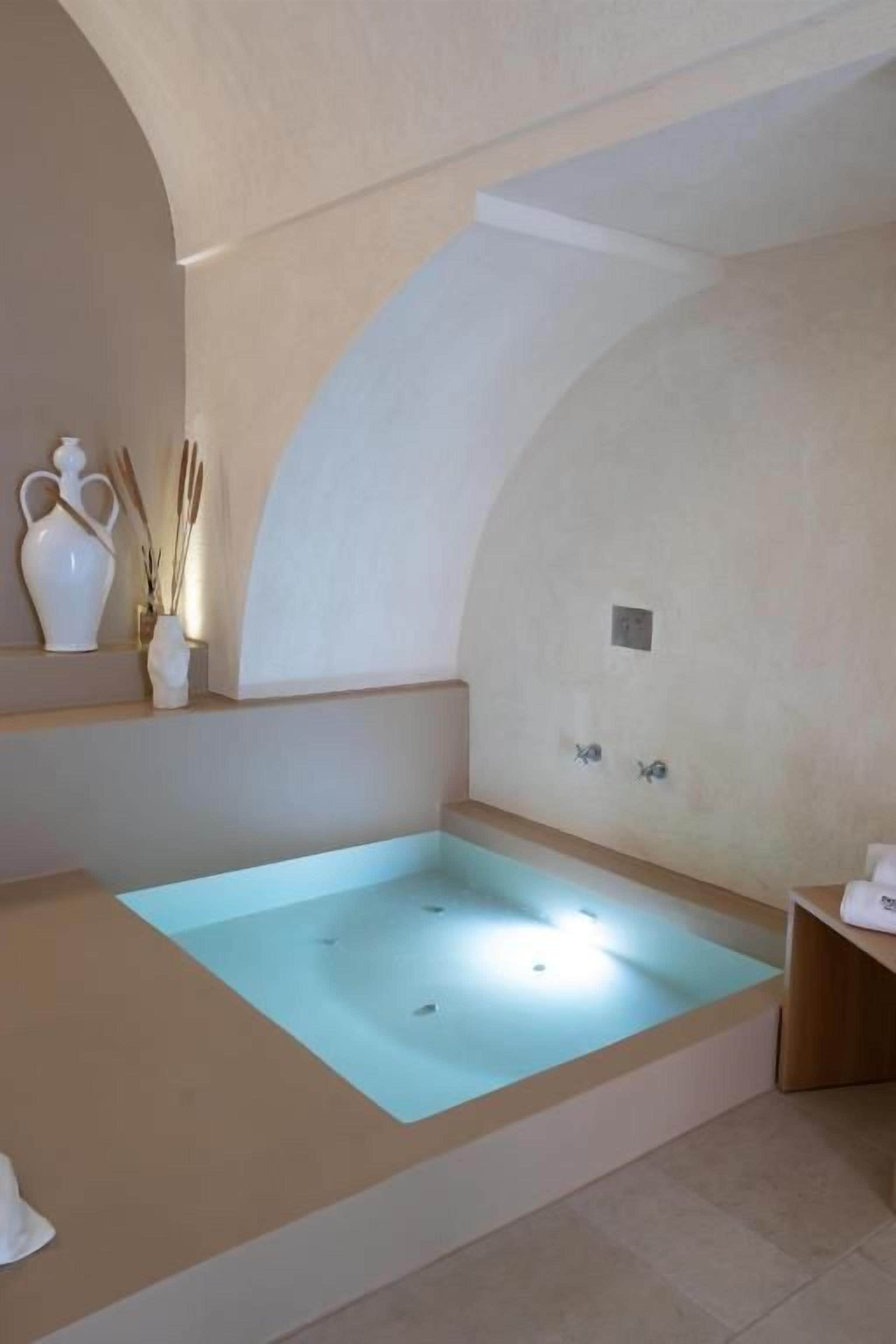 A spa room with a large bathtub, a small shelf with decorative items, and a wall-mounted light fixture.