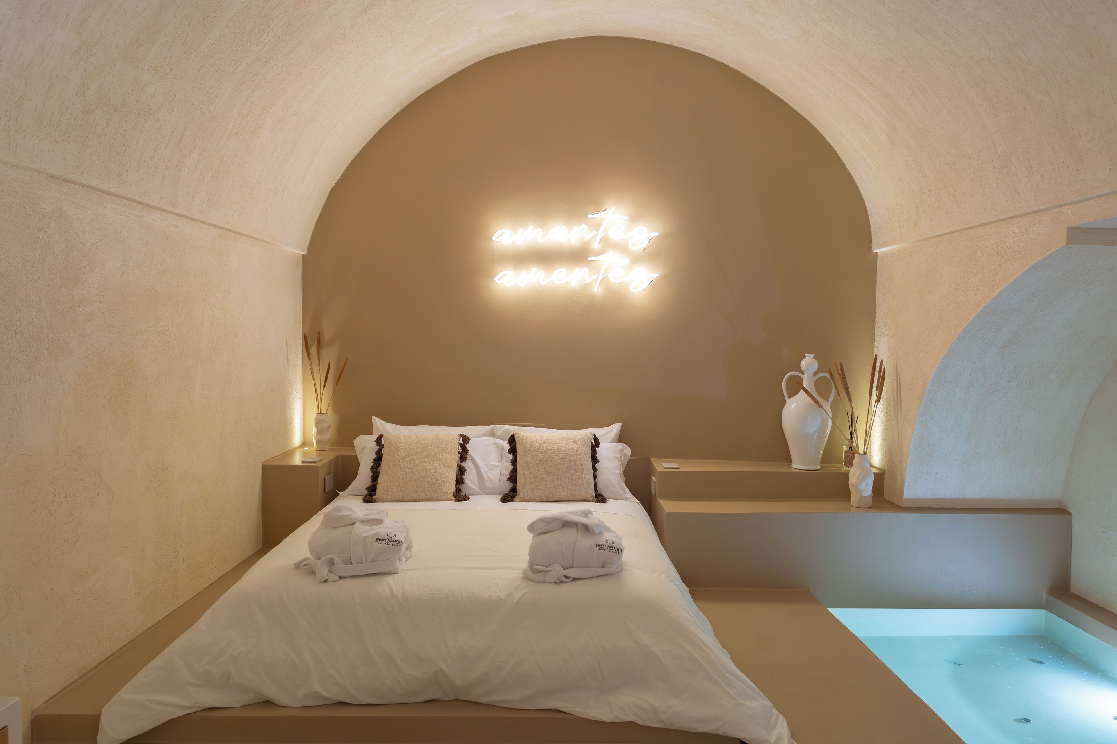 Une chambre d’hôtel dotée d’un grand plafond en arc de cercle, d’un lit avec une literie blanche, de deux serviettes, d’une table de chevet avec un vase et d’un luminaire fixé au mur.