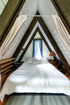 Ein Schlafzimmer mit schräg steilender Decke, einem Bett, einem Nachttisch, einer Lampe und einem Fenster mit Vorhängen.