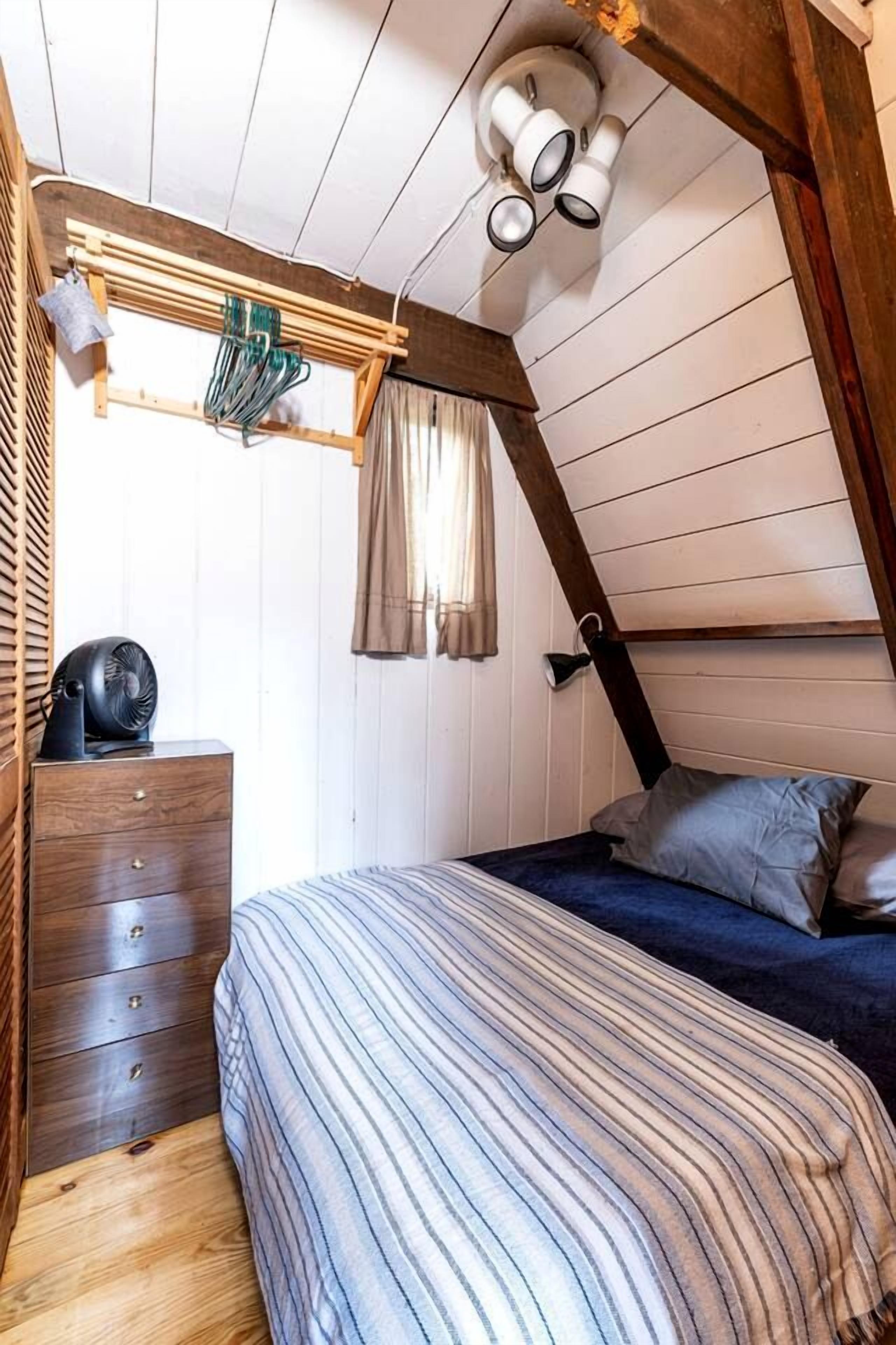 Ein Schlafzimmer mit schrägen Wänden, einem Bett mit gestreifter Decke, einer hölzernen Kommode und einem Deckenventilator.