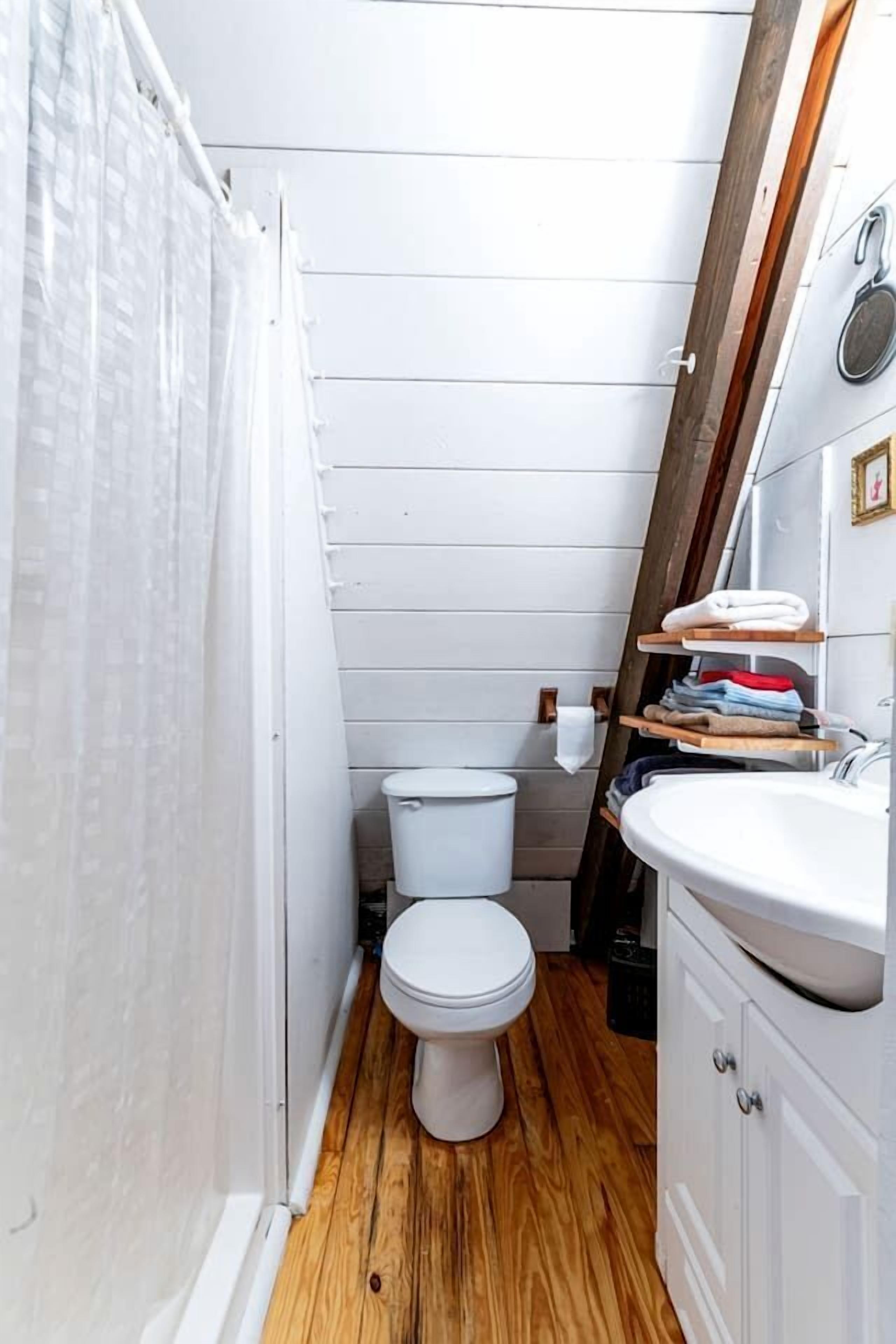 Un baño pequeño con inodoro blanco, suelo de madera, lavamanos blanco y estante de madera con toallas.