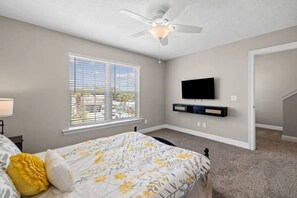 Ein Schlafzimmer mit einem Bett, einem Deckenventilator, einem an der Wand montierten Fernseher und einem Fenster mit Jalousien.