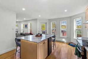A spacious kitchen with a central island, wooden cabinets, and a dining area with a gray sofa. - BLVD QV Lake House  4BR  Backyard  Pets  Parking (St. John's)