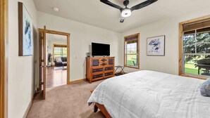 Un dormitorio con una cama, ventilador de techo, televisor sobre una cómoda de madera y ventana con vistas al exterior.