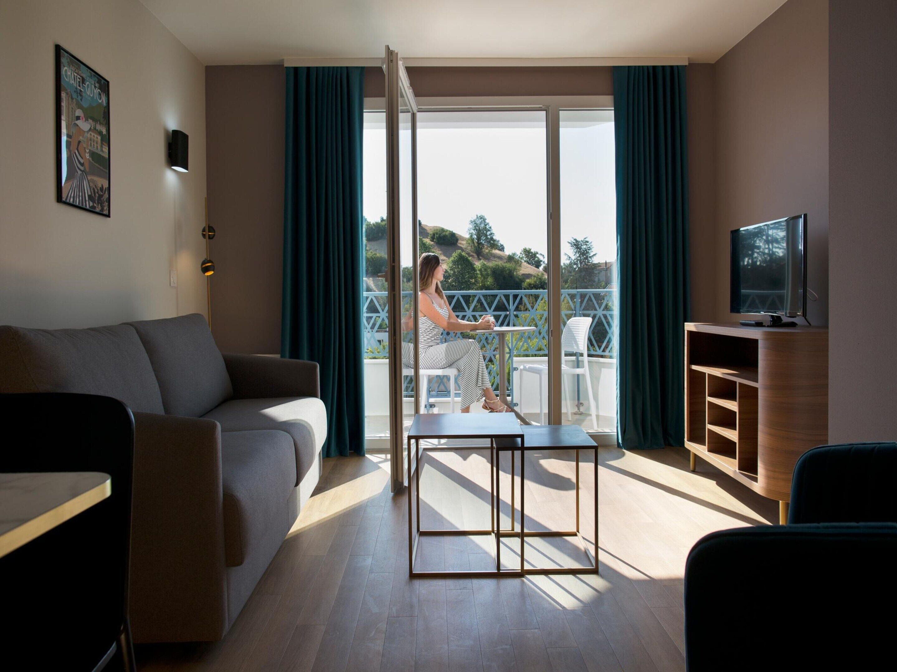 A modern hotel room with a sofa, a TV, a balcony with a table and chairs, and a large window.