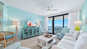 A living room with a white sofa, a wicker chair, a blue armchair, a white coffee table, and a teal cabinet. - Beachfront Corner Views, Indoor & Outdoor Pools, Walk to Dining | BEACHBALL PROPERTIES (Orange Beach)