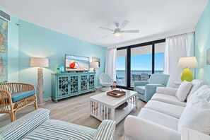A living room with a white sofa, a wicker chair, a blue armchair, a white coffee table, and a teal cabinet. - Beachfront Corner Views, Indoor & Outdoor Pools, Walk to Dining | BEACHBALL PROPERTIES (Orange Beach)