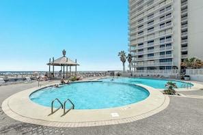 A pool area with a gazebo, lounge chairs, and a tall building in the background. - Beachfront Corner Views, Indoor & Outdoor Pools, Walk to Dining | BEACHBALL PROPERTIES (Orange Beach)