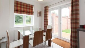 Un coin repas avec une table en verre, des chaises, un radiateur et une vue sur l’extérieur à travers une fenêtre ornée de rideaux.
