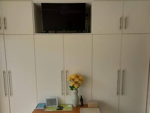 A room with a TV mounted on a cabinet, a vase with yellow flowers, and a small table with a sign and a remote control.