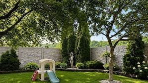 A backyard with a play area, including a slide and climbing structure, surrounded by a stone wall and lush greenery.