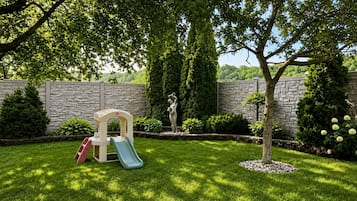 Un jardin avec un espace de jeux, comprenant un toboggan et une structure d’escalade, le tout entouré d’un mur de pierres et d’une végétation luxuriante.