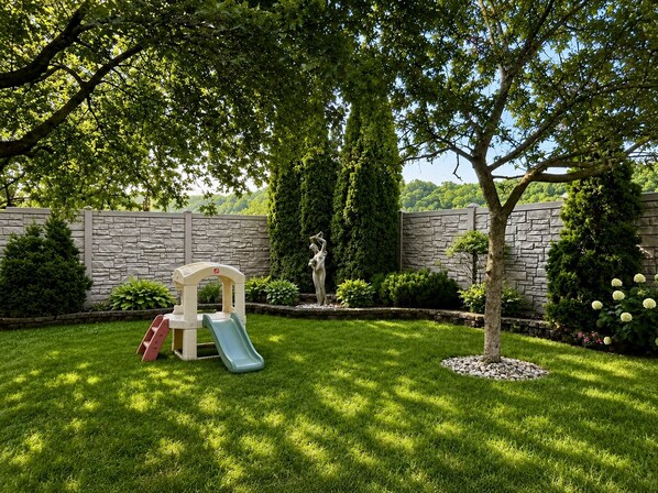 A backyard with a play area, including a slide and climbing structure, surrounded by a stone wall and lush greenery.