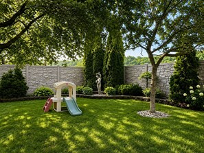 Un patio trasero con zona de juegos, que incluye un tobogán y una estructura para escalar, rodeado por un muro de piedra y abundante vegetación.