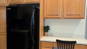 A kitchen with wooden cabinets, a black refrigerator, a white countertop, a black chair, and a small plant.
