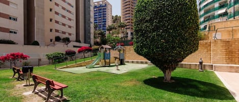 Une cour avec un espace de jeux, des bancs et une haie bien entretenue.