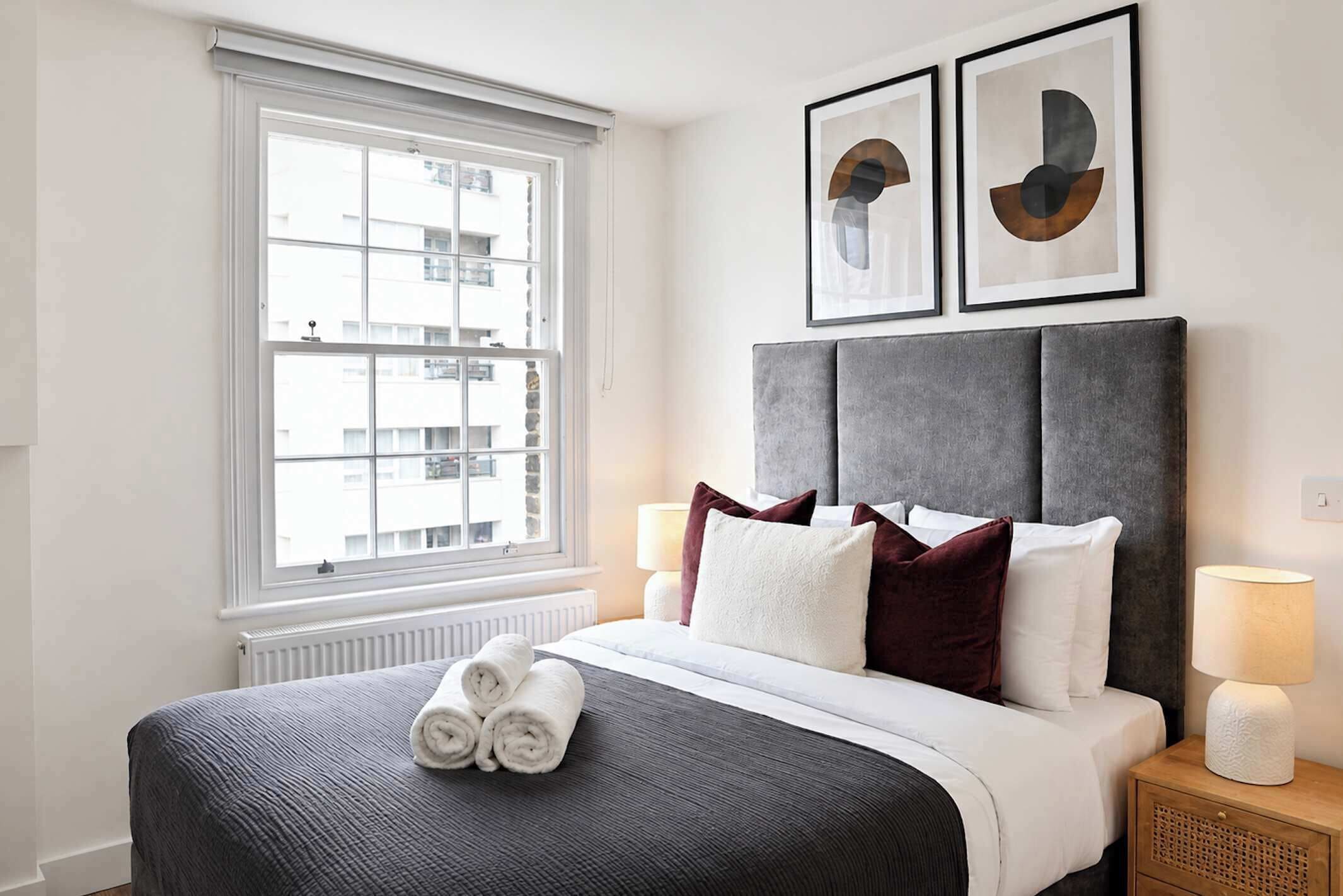 A hotel room with a large window, a bed with rolled towels, a grey headboard, two framed abstract artworks, and a bedside lamp.