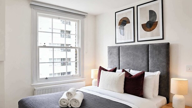 Una habitació d'hotel amb una finestra gran, un llit amb tovalloles arremolinades, un capçal de llit gris, dues obres d'art abstractes emmarcades i una làmpada de nit.