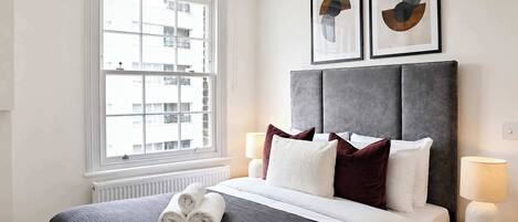 Una habitación de hotel con un ventanal grande, una cama con toallas enrolladas, un cabecero gris, dos obras de arte abstracto enmarcadas y una lámpara de noche.