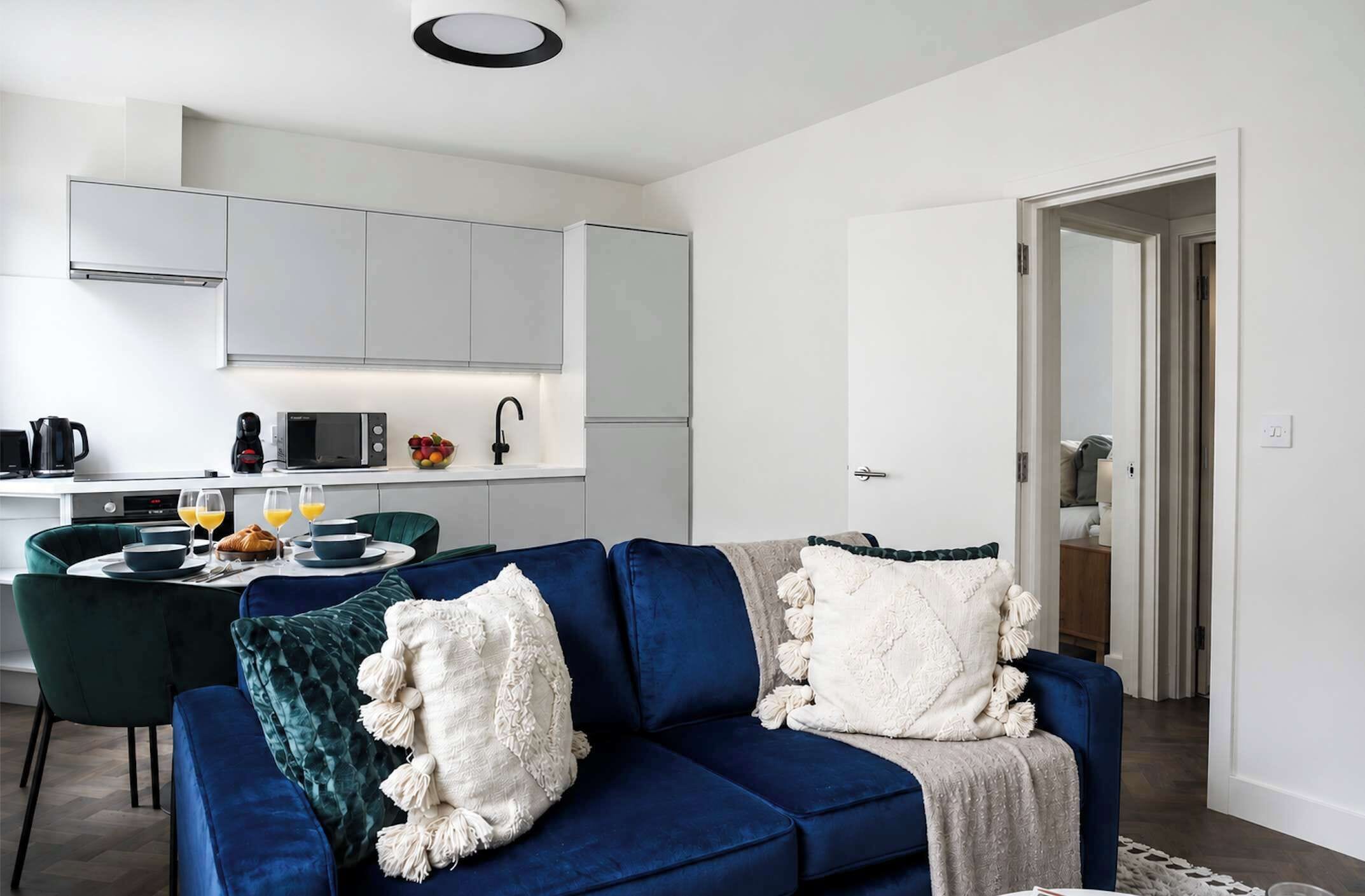 A modern living room with a blue sofa, a dining area with a table and chairs, and a kitchen with white cabinets and appliances.