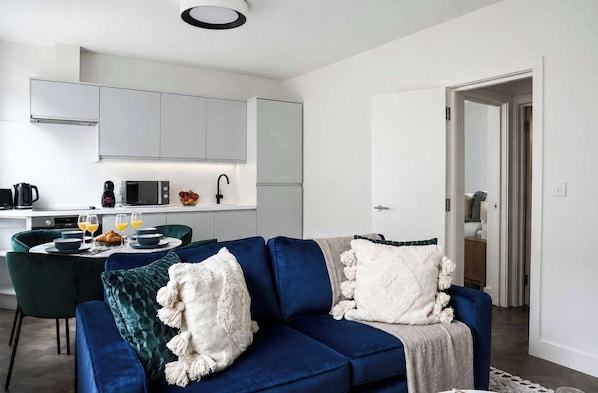 Ein modernes Wohnzimmer mit einem blauen Sofa, einem Essbereich mit Tisch und Stühlen sowie einer Küche mit weißen Schränken und Geräten.