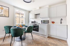 Uma cozinha moderna com armários brancos, uma área de refeições com cadeiras verdes e um chão de madeira em padrão herringbone.