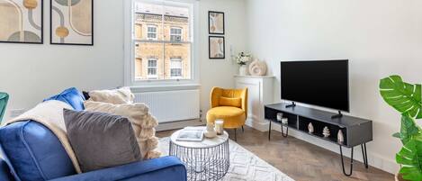Una sala de estar moderna con un sofá azul, un sillón amarillo, un televisor de pantalla plana y un ventanal con vistas a un edificio de ladrillo.
