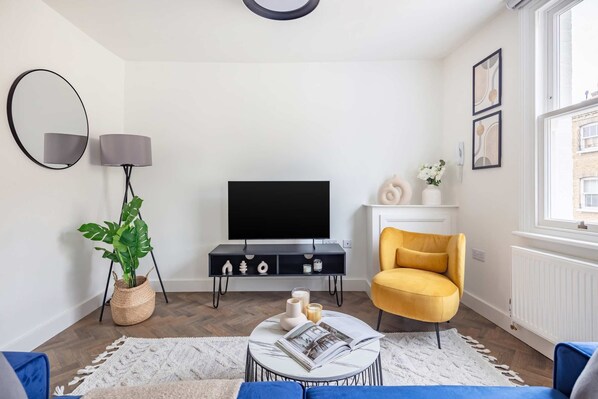 Uma sala de estar moderna com televisão de tela plana, uma poltrona amarela, uma mesa de centro redonda com uma revista e um vaso de planta.