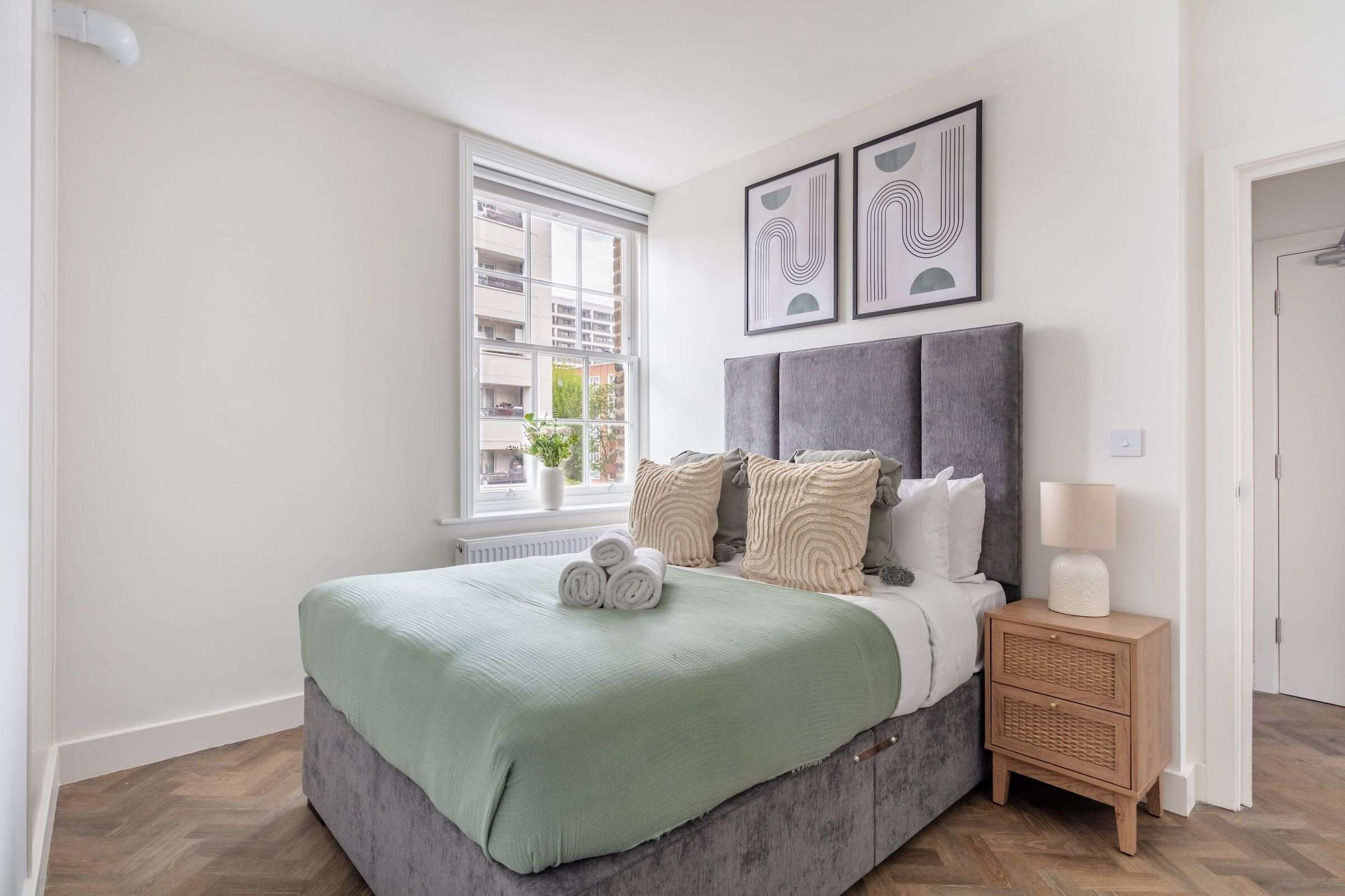A bedroom with a bed, a bedside table, a lamp, a window, and artwork on the wall.