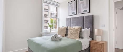 Un dormitorio con cama, mesita de noche, lámpara, ventana y una obra de arte en la pared.