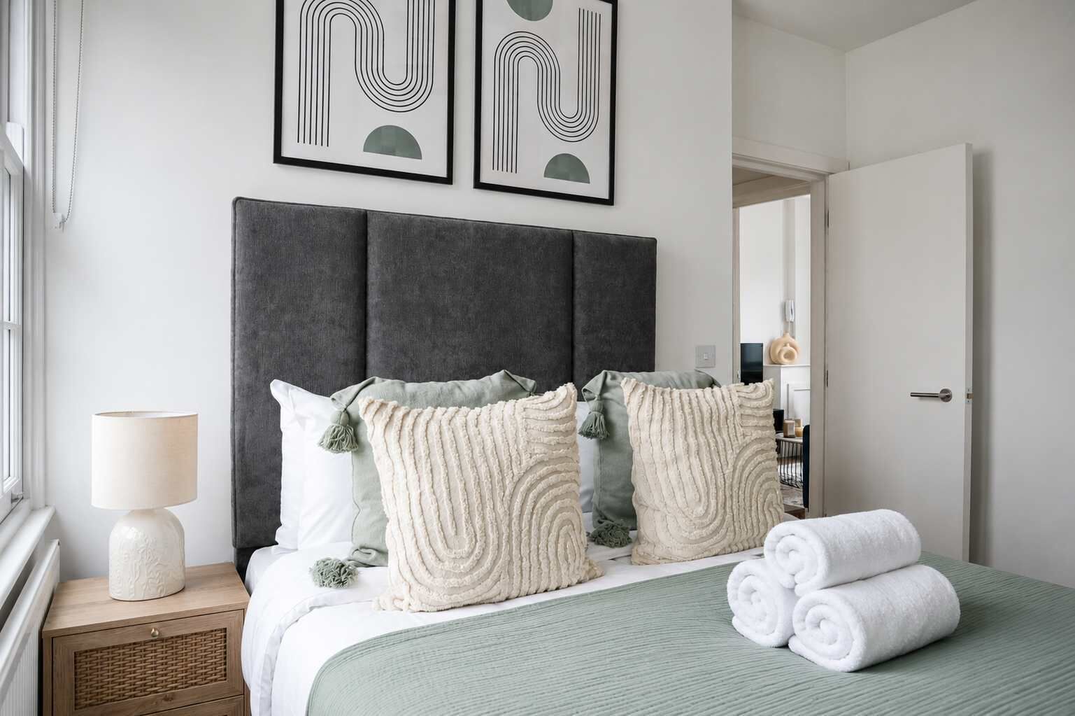 Una cama bien hecha con toallas enrolladas, una lámpara de noche y almohadones decorativos.