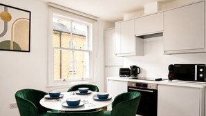 Une cuisine compacte avec des meubles blancs, un micro-ondes noir et une machine à café. On y trouve une table à manger avec des bols bleus et une chaise verte.