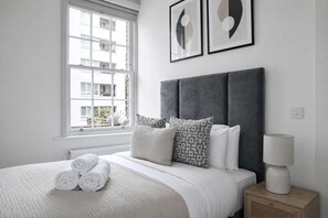 Ein Schlafzimmer mit Bett, Kissen, einer Nachttischlampe und einem Fenster mit Blick auf Gebäude.