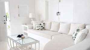 Una sala de estar blanca con una mesa de centro de vidrio, un sofá con cojines estampados, una mesita auxiliar y una puerta.