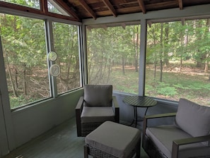 Une véranda couverte avec deux fauteuils confortables et une petite table, de grandes fenêtres offrant une vue sur une zone boisée.