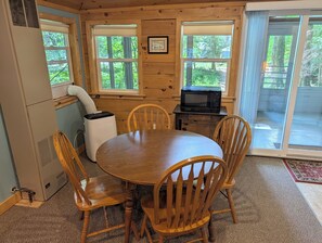 Ambiente interno in legno con un tavolo rotondo, quattro sedie, un forno a microonde e una finestra vista verde.