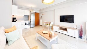 A modern living room with a sofa, coffee table, TV, and kitchen area. - Bright Modern 1BR · 4-min Walk to the Sea · Alimos Riviera (Alimos)